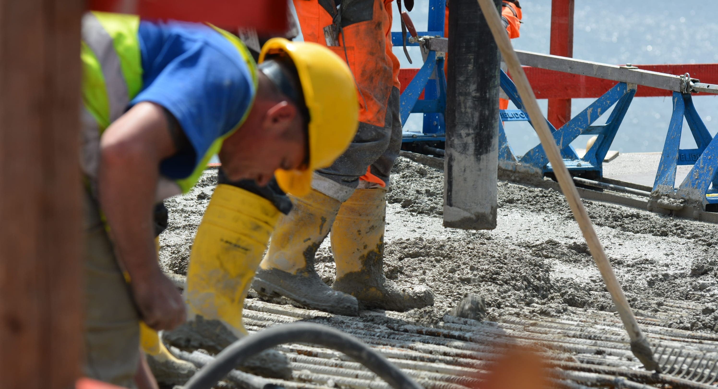 Das Bild zeigt einen Nahbereich einer Baustelle, auf der Beton gegossen wird. Ein Arbeiter mit gelbem Helm und Warnweste beugt sich über die Arbeit, während er mit Werkzeugen hantiert. Der Boden ist mit frischem Beton bedeckt, und gelbe Gummistiefel schützen die Füße der Arbeiter. Im Hintergrund sind rote und blaue Bauelemente zu erkennen, die die Baustelle strukturieren.

                                